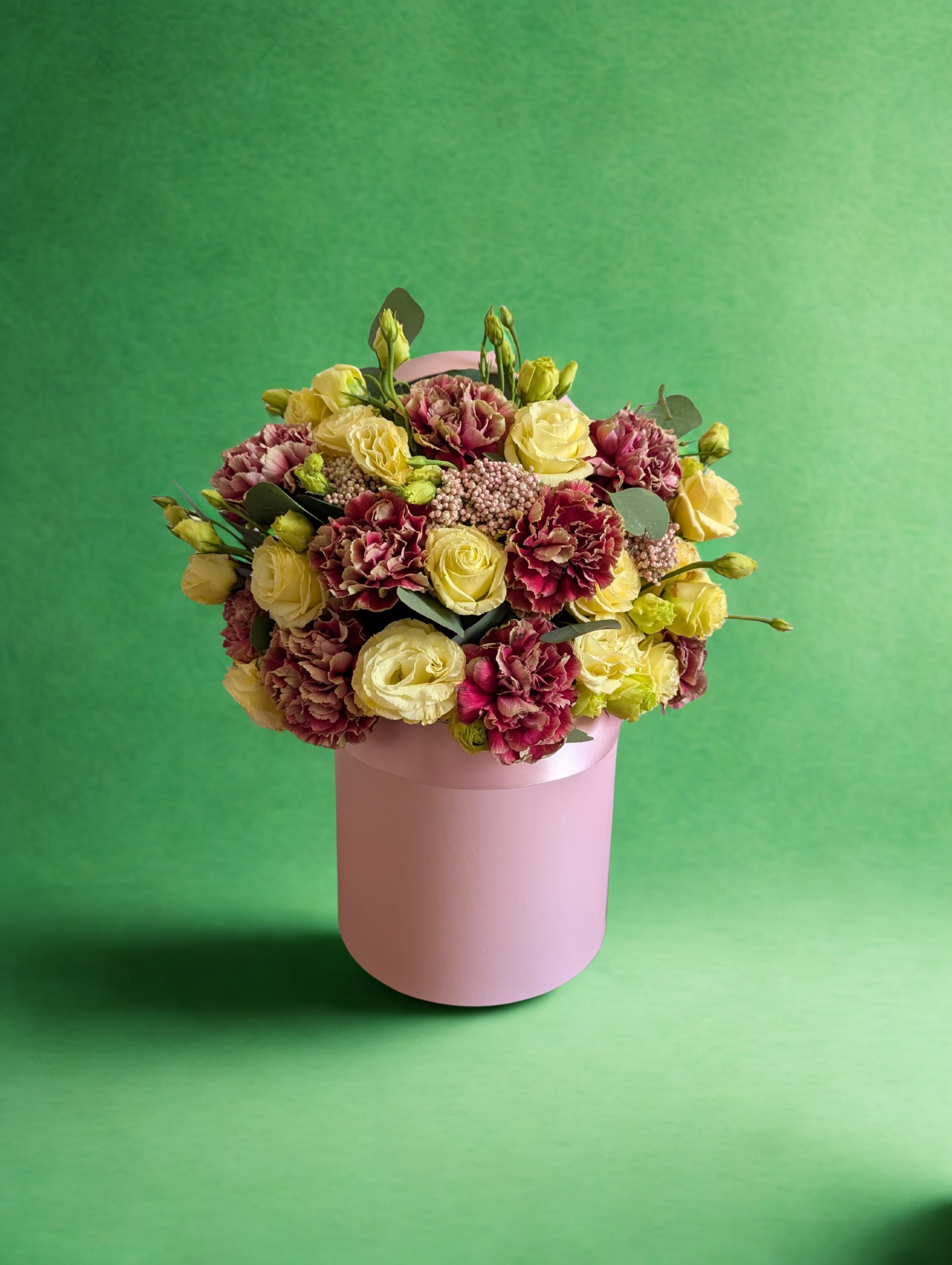 Antique Garden hat box floral arrangement with dusty mauve carnations, soft yellow spray roses in full bloom and bud, and waxflower in a blush pink satin-banded box with silver dollar eucalyptus