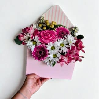 Hand holding a pink envelope box overflowing with pink roses, magenta gerbera daisies, white daisies, and alstroemeria — Irus Floral Desig